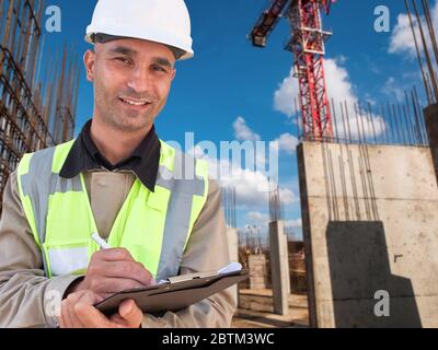 Construction worker in white hardhat stands with clipboard opposite construction site with tower crane at background. Real estate expert, civil engine Stock Photo