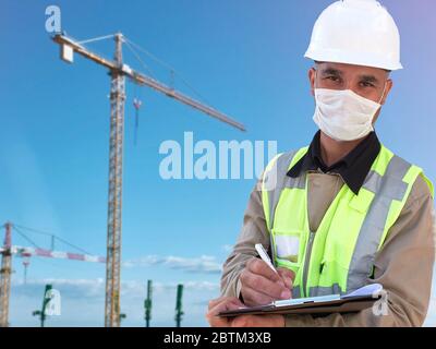 Construction worker in white hardhat and protective mask stands with clipboard opposite construction site with tower cranes at background Stock Photo