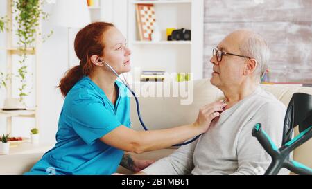 Caucasian female nurse listening to elderly retired man heartbeat in bright and cozy nursing home. Social worker and caregiver Stock Photo