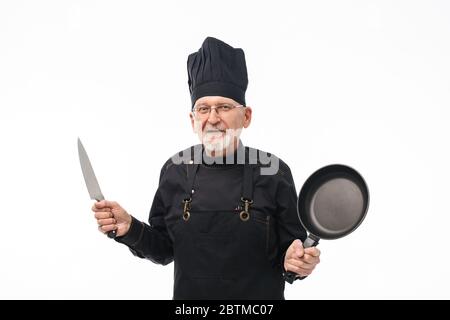 Old cheerful man with gray beard in black apron and cap holding pan and knife in hands while joyfully looking in camera over white background Stock Photo