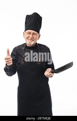 Old attractive smiling man with gray beard in black apron and cap holding pan in hand showing thumb up gesture while dreamily looking in camera over Stock Photo