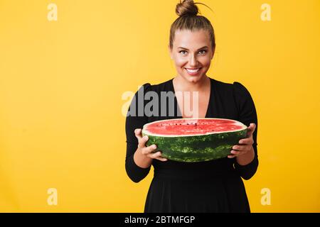 Cheerful girl in black dress joyfully looking in camera while holding half of big watermelon in hands over yellow background Stock Photo