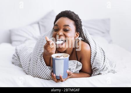 Happy african american girl puts spoon in her mouth and holds bucket of ice cream Stock Photo