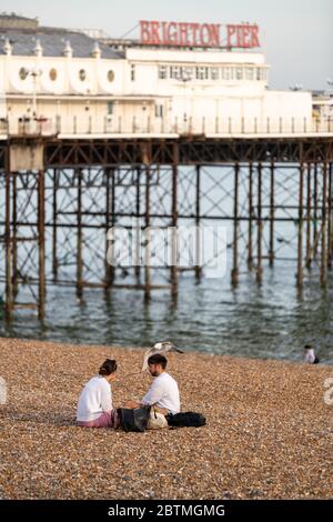 a couple sitting on pebbled beach in front of Brighton Palace Pier in lockdown during Covid-19 pandemic Stock Photo