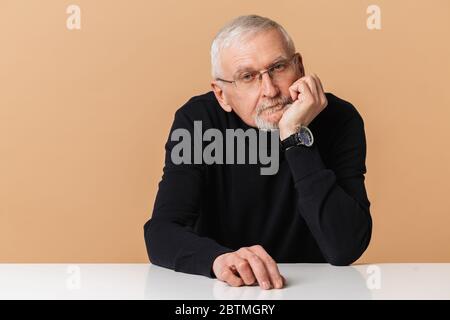 Old attractive man with gray hair and beard in eyeglasses and sweater leaning on hand thoughtfully looking in camera over beige background isolated Stock Photo