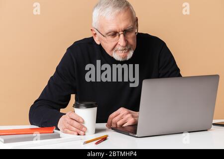 Old handsome man with gray hair and beard in eyeglasses and sweater sitting at the table with books and coffee to go thoughtfully working on laptop Stock Photo