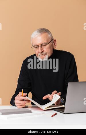 Old handsome man with gray hair and beard in eyeglasses and sweater sitting at the table with laptop thoughtfully writing in notebook over beige Stock Photo