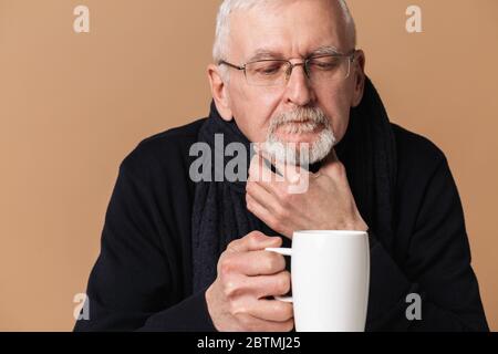 Old sick man with gray hair and beard in eyeglasses and scarf with big cup with tea while holding hand near throat over beige background Stock Photo