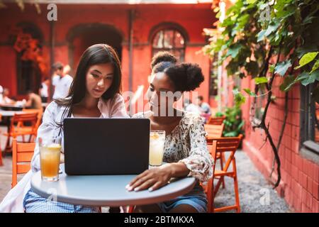 Two beautiful girls thoughtfully using laptop while spending time with cocktails together in cozy courtyard of cafe Stock Photo