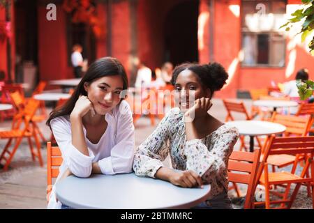 Two beautiful smiling girls leaning on hands dreamily looking in camera while spending time together in courtyard of cafe Stock Photo