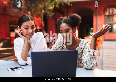 Two beautiful smiling girls working with laptop together while spending time in cozy courtyard of cafe Stock Photo