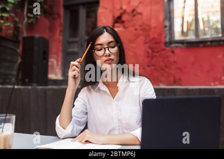 Beautiful girl with dark hair in white shirt and eyeglasses thoughtfully working with laptop in courtyard of cafe Stock Photo