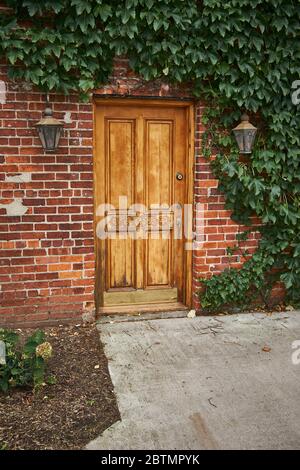 Closed four panel wooden door with ornate carvings surrounded with green ivy opening into brick building. Lights at door, Concrete sidewalk entrance. Stock Photo