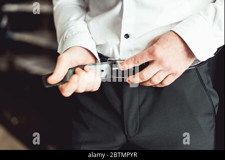 Groom in black pants and a white shirt fastens his leather belt. grooms mourning. Wedding details. Stock Photo