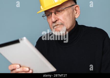 Close up old man with gray beard in eyeglasses and yellow safety helmet thoughtfully using the tablet over blue background isolated Stock Photo