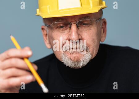 Close up old pensive man with gray beard in eyeglasses and yellow safety helmet thoughtfully looking aside while holding pencil in hand over blue Stock Photo
