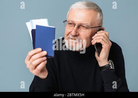 Old handsome smiling man with gray hair and beard in eyeglasses and sweater holding tickets and passports in hand while happily talking on cellphone Stock Photo