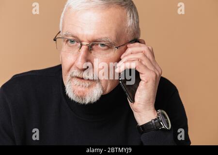 Old attractive man with gray hair and beard in eyeglasses and sweater thoughtfully looking aside while talking on cellphone over beige background Stock Photo