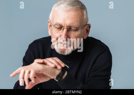 Old handsome man with gray hair and beard in eyeglasses and sweater dreamily looking on hand watch over blue background isolated Stock Photo