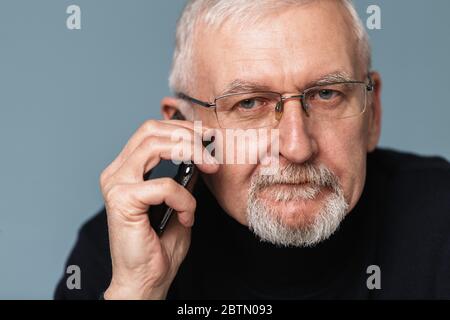 Old attractive man with gray hair and beard in eyeglasses and sweater talking on cellphone while dreamily looking in camera over blue background Stock Photo