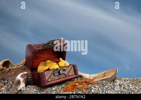 A small treasure chest filled with gold nuggets. Chest is on a sandy beach with driftwood. Background is blue sky with clouds. Stock Photo