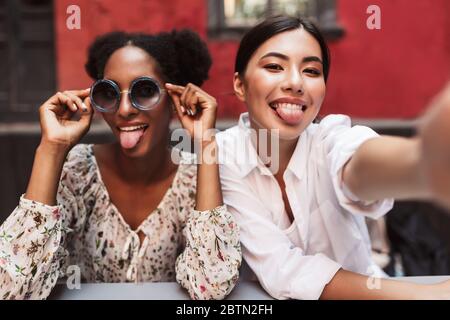Two pretty girls happily showing tongues while taking photos together spending time in courtyard of cafe Stock Photo