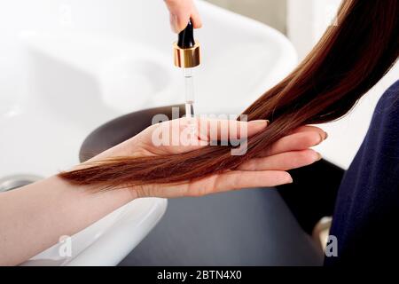 Female hands applying oil on long womans brown hair. Hair care cosmetics, bath beauty spa products. Female hair serum for treatment hair tips against Stock Photo
