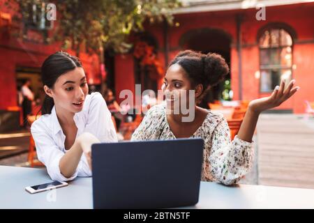 Young pretty emotional ladies working with laptop together while spending time in cozy courtyard of cafe Stock Photo