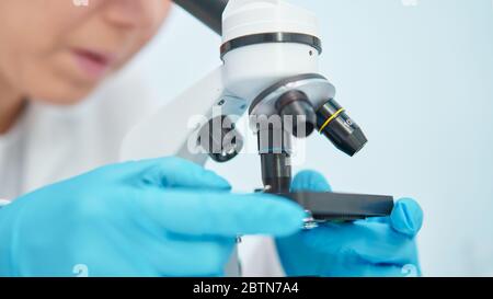 Close-up of female lab technician in blue rubber gloves looking through microscope in laboratory. Stock Photo