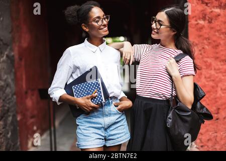 Two beautiful smiling girls in eyeglasses with laptop and backpack happily looking at each other Stock Photo