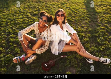 Two pretty smiling girls sitting on grass happily looking in camera while spending time together in city park Stock Photo