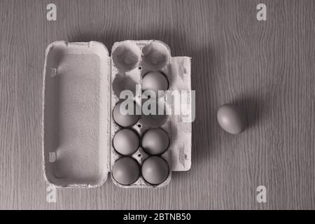 A black-and-white image of chicken eggs lying in a cardboard box, which stands on a wooden table, and one egg lies next to the box.