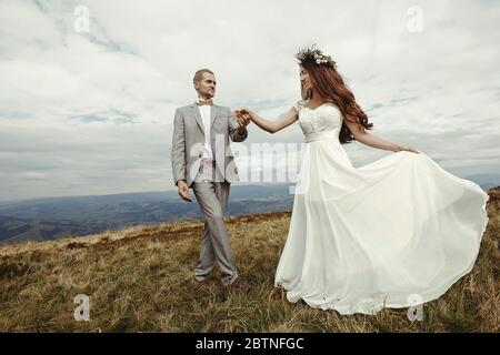 happy gorgeous bride and groom holding hands and dancing  in sun light, boho wedding couple, luxury ceremony at mountains with amazing view Stock Photo