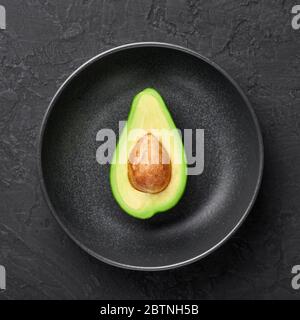 Half Avocado in black rough bowl on dark slate backdrop. Black ceramic dish setting. Stock Photo