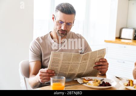 Photo of serious caucasian man in eyeglasses reading newspaper while having breakfast in modern kitchen Stock Photo