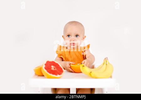 Happy baby sitting in high chair eating fruits in a white kitchen. Healthy nutrition for kids. Children eat oranges and grapefruits. Stock Photo