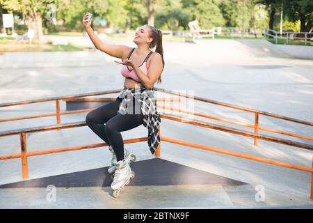 Young pretty smiling plus size woman in pink sporty top,leggings and roller skates leaning on railing happily sending air kiss while taking photo on Stock Photo