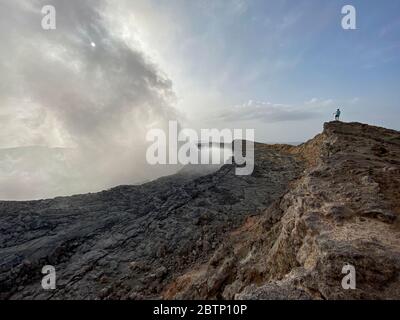 Hiker on rocks looking at smoke coming out from Erta Ale volcano, Danakil Depression, Afar Region, Ethiopia, Africa Stock Photo