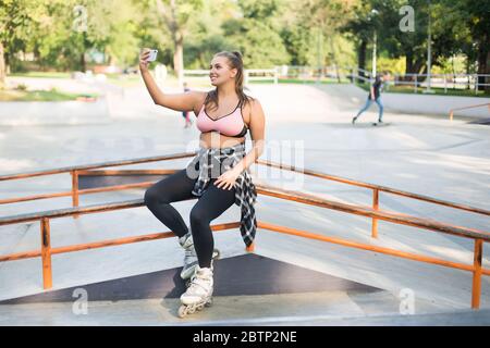 Young beautiful smiling plus size woman in pink sporty top,leggings and roller skates leaning on railing happily taking photo on cellphone while Stock Photo