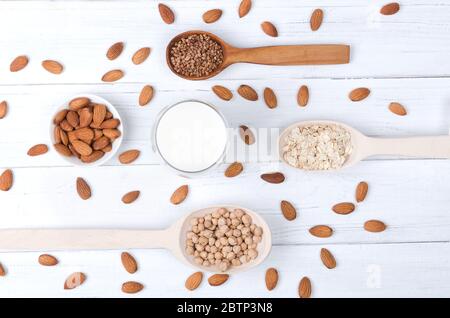 Flatlay view of almond milk in glass with almond seeds, buckweat, oat and wooden spoons on white wooden table Stock Photo