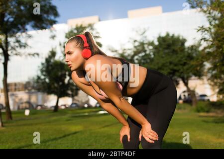 Young tired plus size woman in sporty top and leggings with red headphones thoughtfully looking aside while leaning on knees in city park Stock Photo