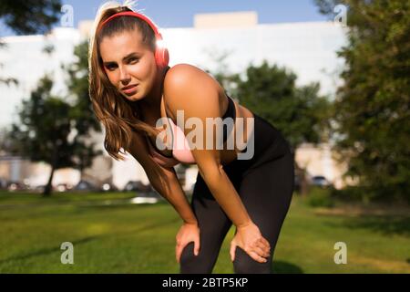 Young tired plus size woman in sporty top and leggings with red headphones thoughtfully looking in camera while leaning on knees in city park Stock Photo