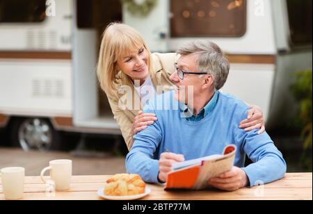Mature woman hugging her husband while he solving crossword during breakfast at campsite Stock Photo