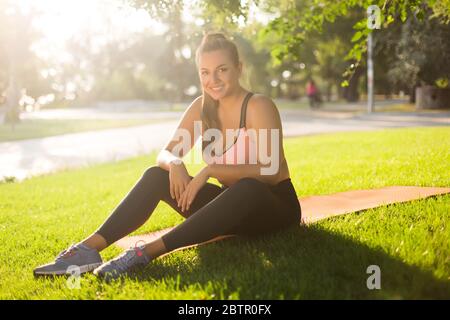 Pretty plus size girl in sporty top and leggings happily doing sport  exercises on orange yoga mat while spending time on green grass in park  Stock Photo