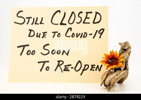 Orange gazania daisy on driftwood next to handwritten STILL CLOSED DUE TO COVID 19, TOO SOON TO RE-OPEN sign Stock Photo