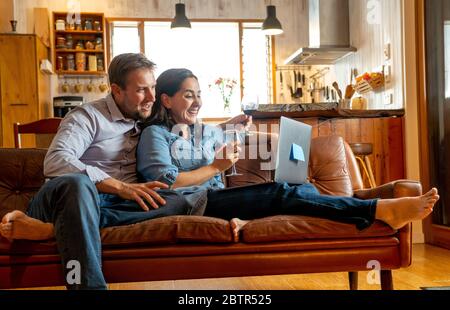 Life at home image of Happy couple video calling friends and family using laptop. Man and woman online chatting cheering with wine in virtual celebrat Stock Photo