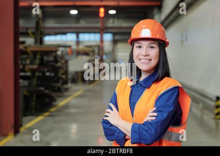 Portrait of smiling confident young Asian factory engineer in orange hardhat and vest standing in workshop Stock Photo