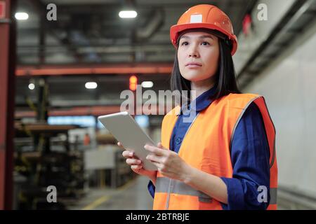 Pensive Asian female process engineer in work helmet and safety vest using digital tablet at factory Stock Photo
