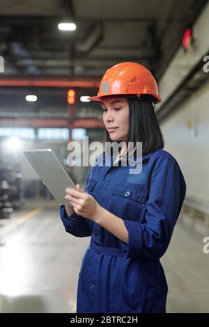 Serious young Asian female engineer in overall analyzing industrial processes with tablet app at factory Stock Photo
