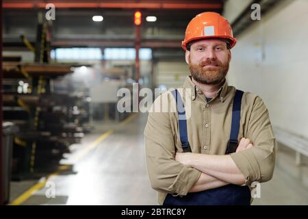 Portrait of content brutal bearded factory man in orange hardhat standing with crossed arms in workshop Stock Photo